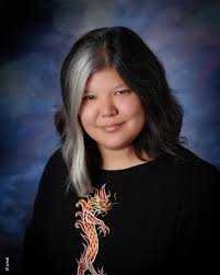 Author Julie Kagawa with medium length dark hair and a bold white streak smiles softly, wearing a black shirt with an orange yellow dragon, posed before a blue purple backdrop inspired by The Iron King Special Edition (The Iron Fey) by Julie Kagawa.