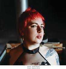 Katee Robert with short, vivid red hair and a nose piercing faces the camera, evoking Wicked Villains vibes in a black halter top. They flaunt a shoulder tattoo and hold "Desperate Measures (Deluxe Edition)" by Katee Robert in a dimly lit room.