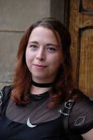 Author Marianne Gordon with long, reddish-brown hair stands in front of a wooden door, smiling softly at the camera and holding "The Gilded Crown" (Raven's Trade) by Marianne Gordon (Paperback).