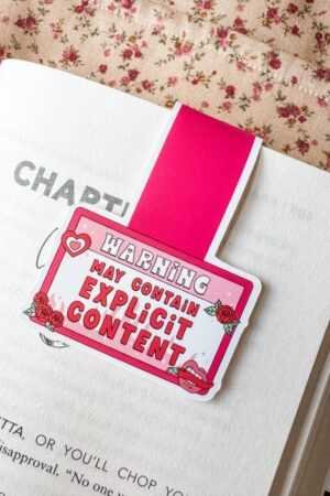 The Magnetic Bookmark Reader Gift Explicit Content, in pink with the phrase "Warning: May Contain Explicit Content," sits on an open book against a floral fabric background—perfect for fun loving readers.