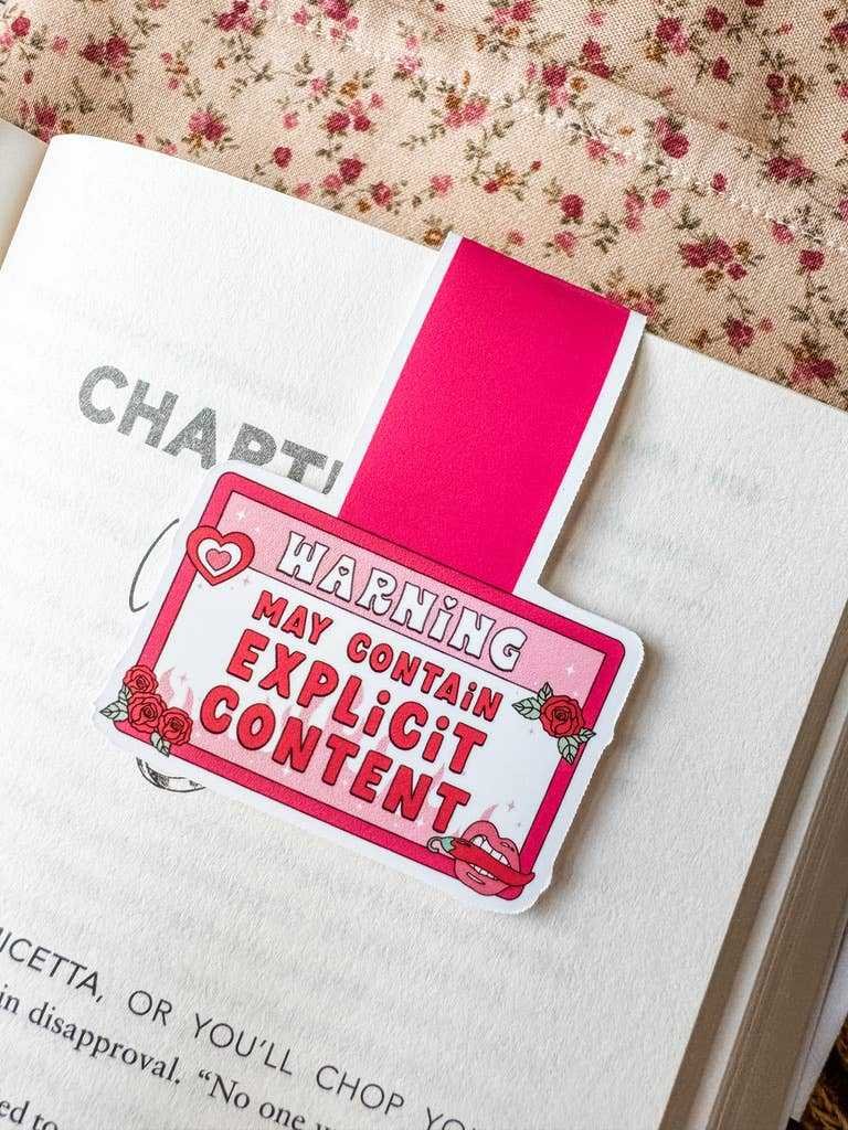 The Magnetic Bookmark Reader Gift Explicit Content, in pink with the phrase "Warning: May Contain Explicit Content," sits on an open book against a floral fabric background—perfect for fun loving readers.