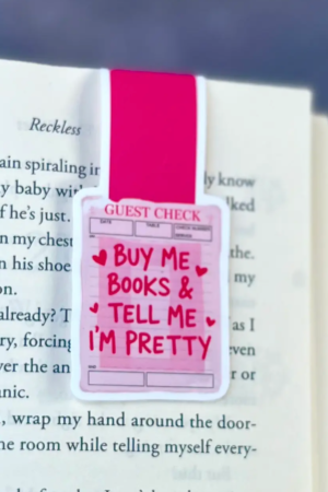 The Magnetic Bookmark Buy Me Books and Tell Me I’m Pretty is a pink and white guest check shaped bookmark clipped to an open book, featuring bold red text and heart accents.
