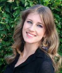 Author Laura Thalassa with long, wavy blonde hair in a black top and large earrings smiles at the camera before a leafy backdrop, her style reminiscent of “Dark Harmony by Laura Thalassa (The Bargainers, 4 of 4).”.