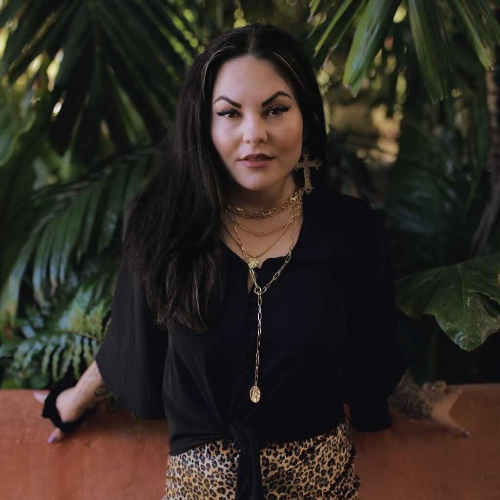 Author Scarlett St. Clair with long dark hair stands before lush leaves, her bold style in black top, layered gold necklaces, and leopard pants echoing the fierce heroines from "Terror at the Gates (Deluxe Edition) - (Blood of Lilith)" by Scarlett St. Clair.