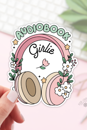 A hand displays the "Audiobook Girlie Stickers / Bookish Stickers / Book Lover" sticker—pink and beige headphones with flowers, "Girlie" center, AUDIOBOOK above. Background: blurred keyboard, notepad, glasses, and greenery. Perfect for book lovers!.