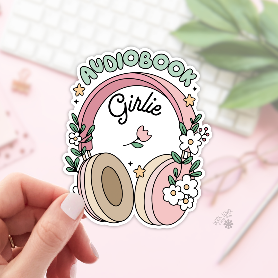 A hand displays the "Audiobook Girlie Stickers / Bookish Stickers / Book Lover" sticker—pink and beige headphones with flowers, "Girlie" center, AUDIOBOOK above. Background: blurred keyboard, notepad, glasses, and greenery. Perfect for book lovers!.