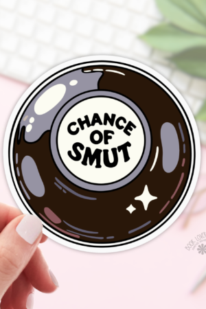 A hand holds the Chance of Smut Stickers, a bookish sticker featuring a Magic 8 Ball design with "CHANCE OF SMUT" in the center. Shown on a pink background with a keyboard, glasses, and plant—ideal for any book lover gift collection.