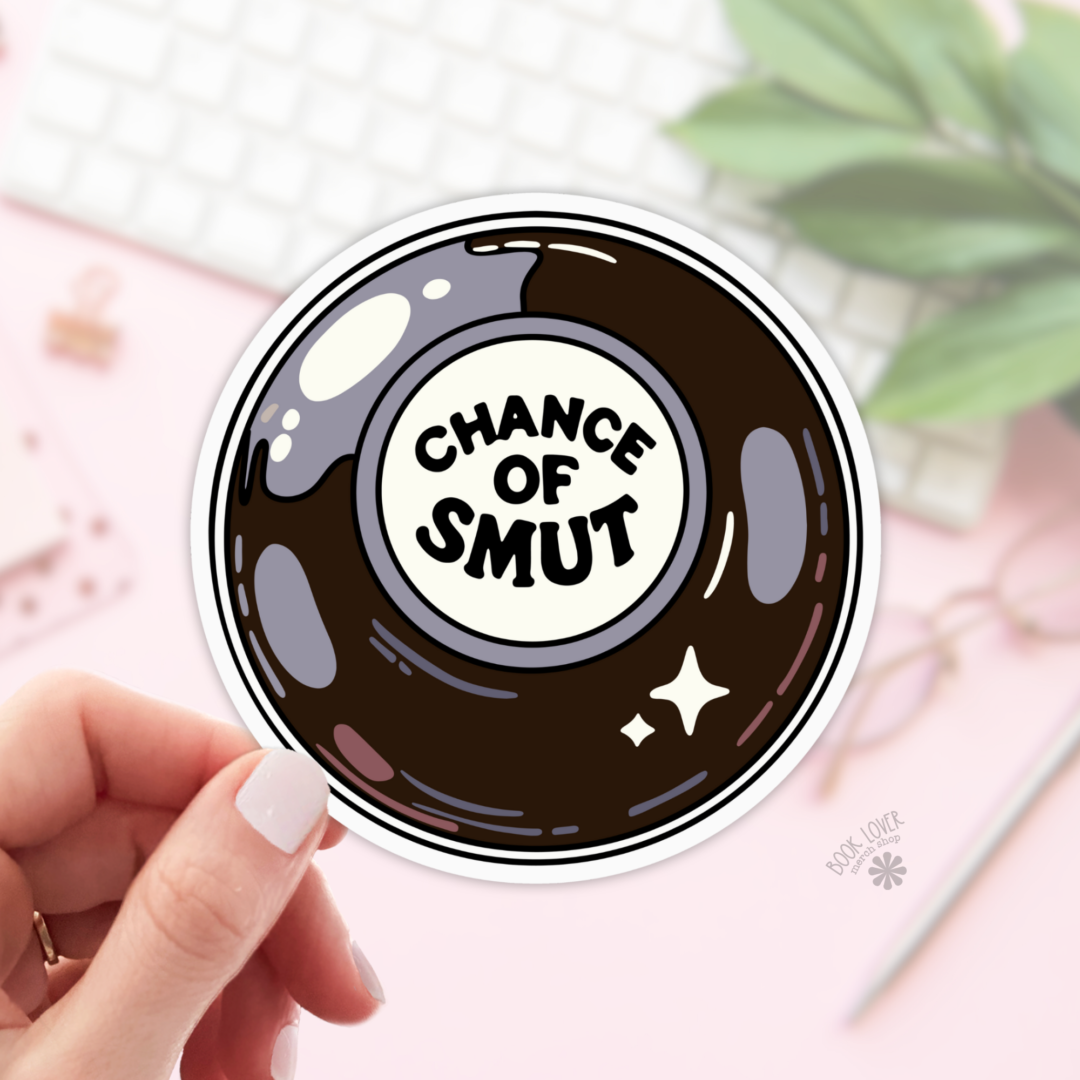 A hand holds the Chance of Smut Stickers, a bookish sticker featuring a Magic 8 Ball design with "CHANCE OF SMUT" in the center. Shown on a pink background with a keyboard, glasses, and plant—ideal for any book lover gift collection.