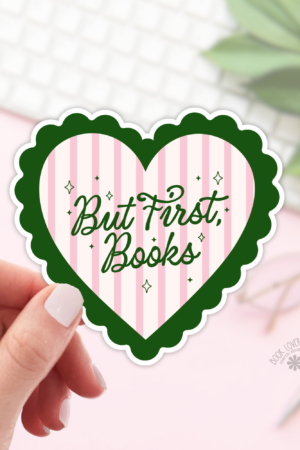 A hand holds a heart-shaped "But First Books" Sticker with a green border and pink stripes from the But First Books Stickers collection. A blurred desk with a keyboard, glasses, and notebook is in the background—ideal for any book lover.