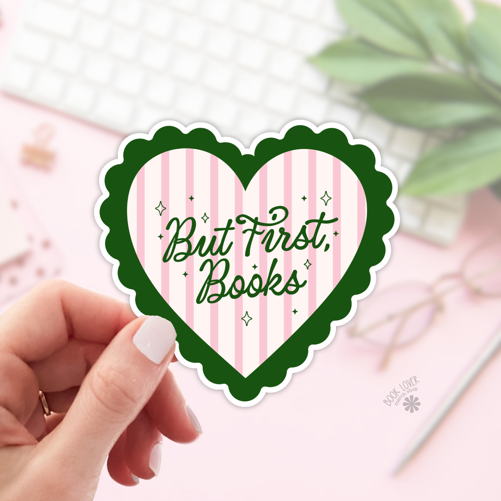 A hand holds a heart-shaped "But First Books" Sticker with a green border and pink stripes from the But First Books Stickers collection. A blurred desk with a keyboard, glasses, and notebook is in the background—ideal for any book lover.
