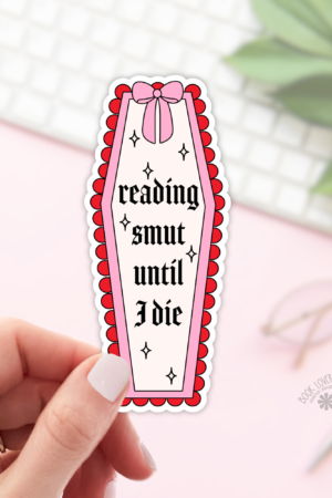 A hand holds the Reading Smut Until I Die Sticker, a coffin-shaped bookish sticker with bold black text, pink border, red scalloped edge, and bow. A keyboard, glasses, and plant are blurred in the background.