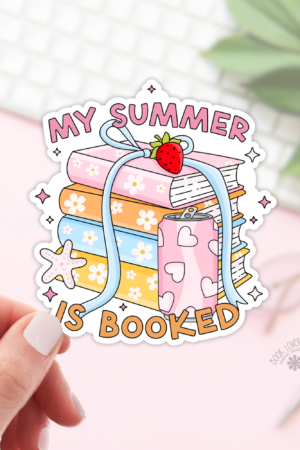 A hand holds a My Summer is Booked Sticker, featuring a stack of colorful books, flowers, a strawberry, and a starfish. Set on a soft pink background with green leaves and keyboard—perfect bookish stickers for any book lover.