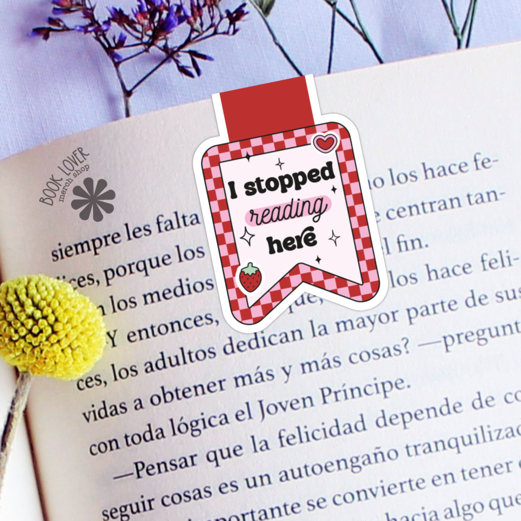 An open book with the "I Stopped Reading Here Strawberry Magnetic Bookmark" lies on a table beside yellow and purple flowers.