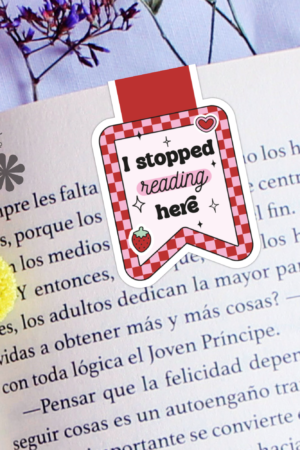 An open book with the "I Stopped Reading Here Strawberry Magnetic Bookmark" lies on a table beside yellow and purple flowers.