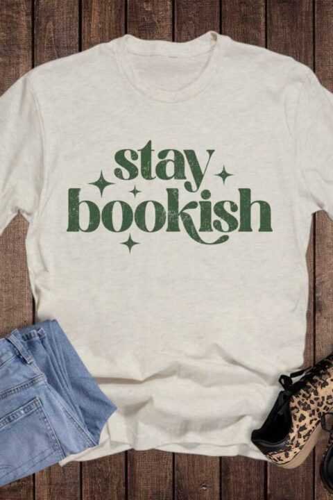 The Reading Books Stay Bookish Graphic Tee, a cream-colored shirt with green "stay bookish" text, is displayed on a wooden floor beside blue jeans and leopard print sneakers.
